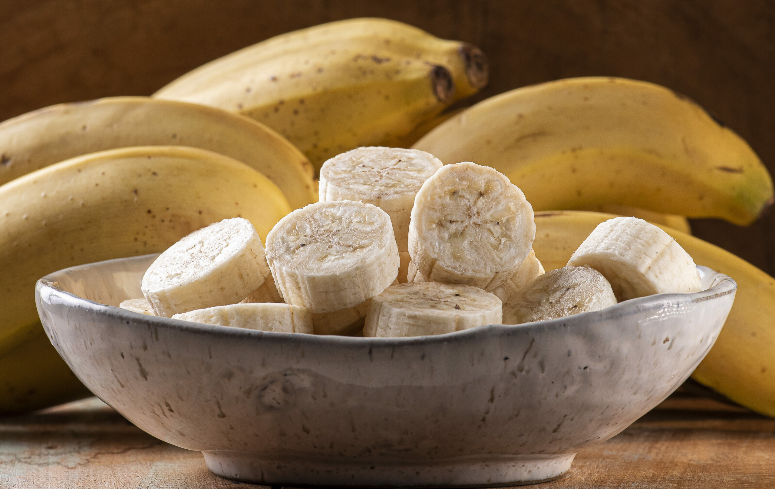 Na foto: Um prato com bananas cortadas em rodelas na frente de cachos de bananas inteiros.